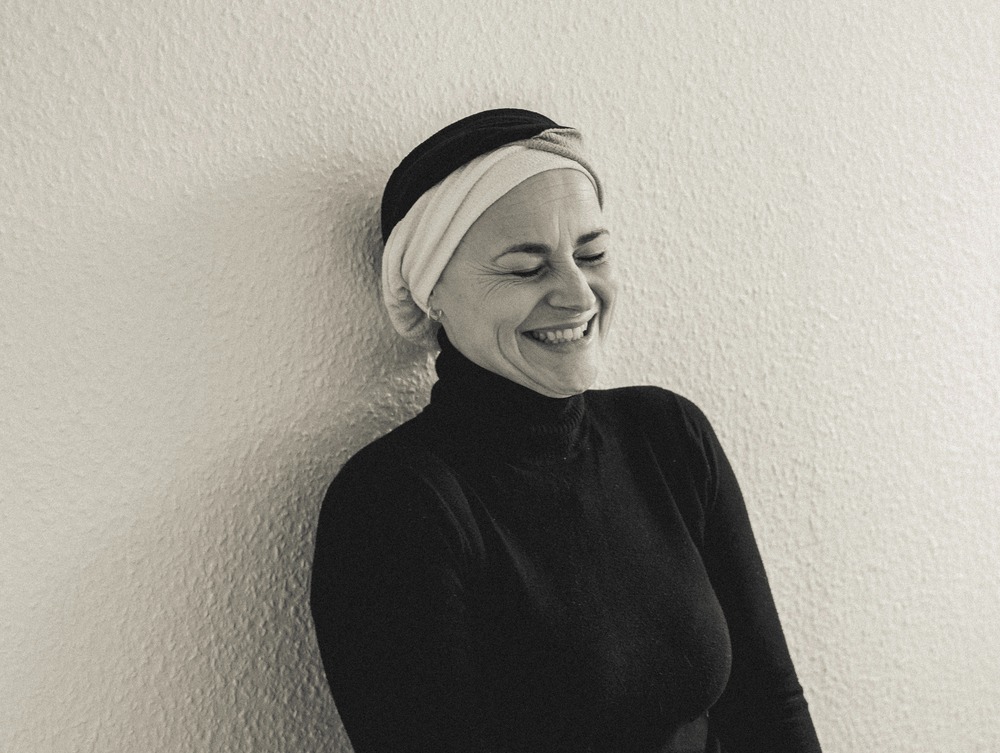 Gudrun læner sig op ad en mur og smiler med en turban om hovedet.