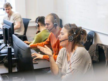 Kvinder sidder og taler i headset foran en computerskærm