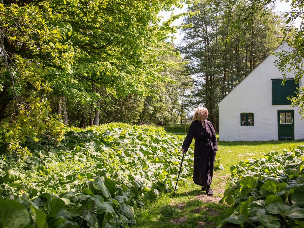 Ulla Koppel stå i haven ved sommerhuset i en kåbe og støtter sig til en stok.