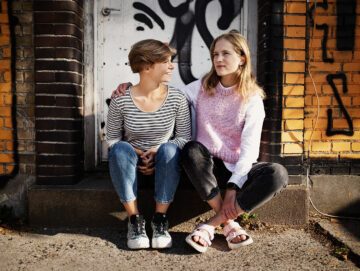 Veninderne Emma Matzen og Mette Grout sidder på et dørtrin, der er graffiti på døren bag dem.