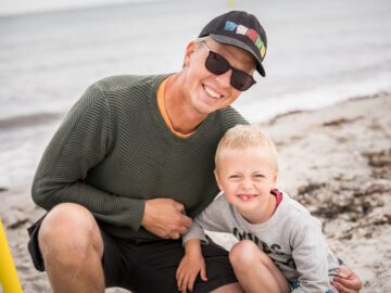 Mand med kasket og solbriller sidder på hug med armen om sin søn på stranden