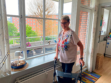 Margit står med sin rollator foran vinduet i sin lejlighed